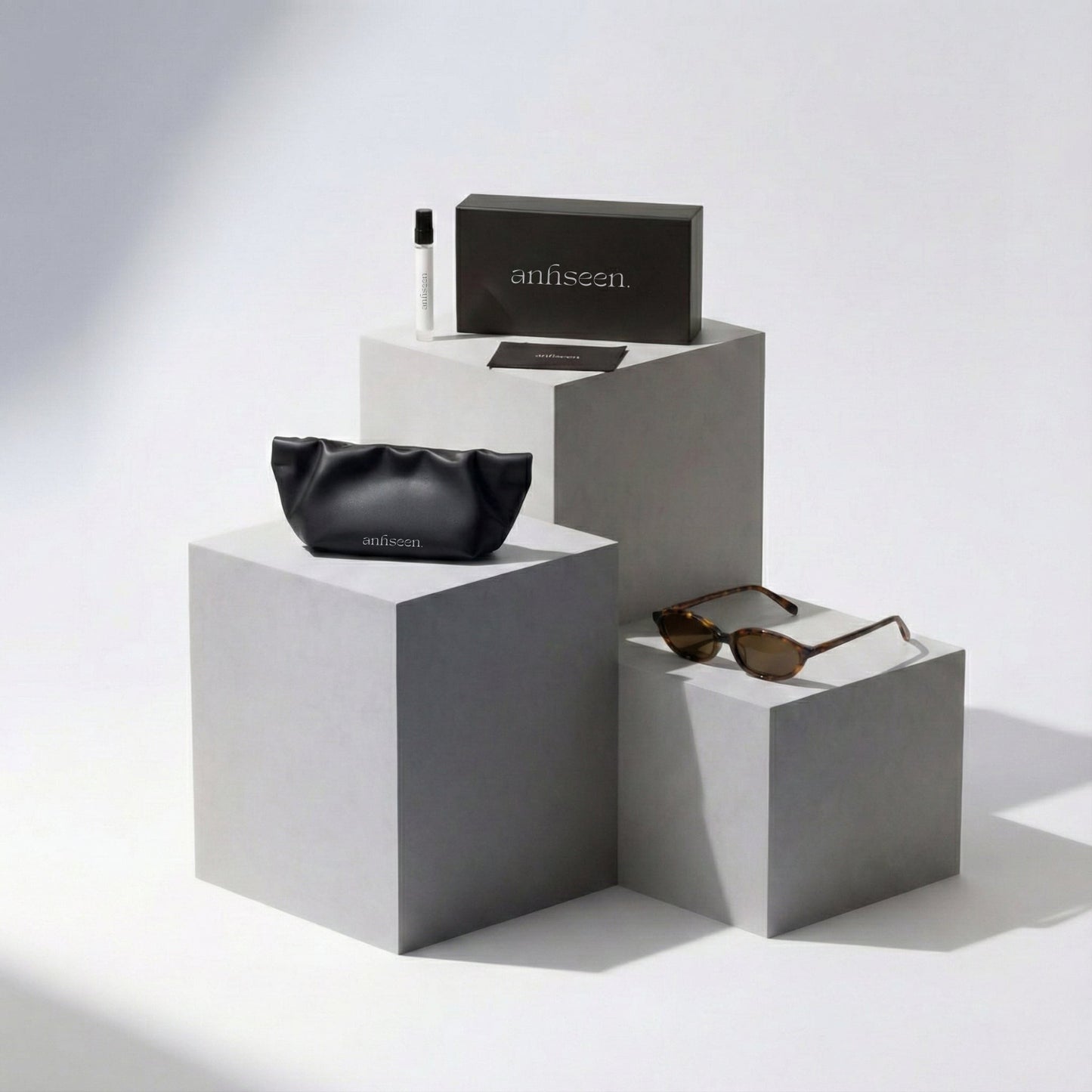 Black pouch, sunglasses, and box on gray cubes with 'anhseen' branding against a white background